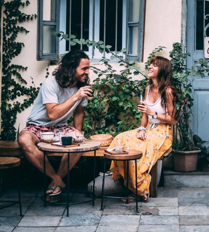 A man and woman sit outside at a small table, enjoying drinks and sharing a laugh. They are surrounded by plants and a building with blue doors and windows.