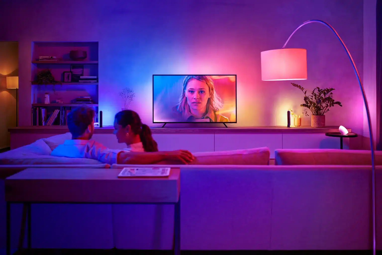 A couple sits on a couch in a dimly lit living room, watching a TV that displays a woman's face. The room is illuminated with colorful ambient lighting.