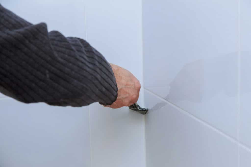 The worker repairing tiler on the wall with trowel, removes white silicone of laying tiles and